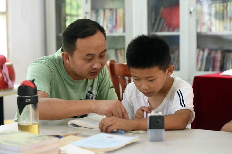 7月22日，在枞阳县横埠镇方正小学安置点“爱心教室”里，方正小学老师操磊（左）指导小朋友写暑假作业。